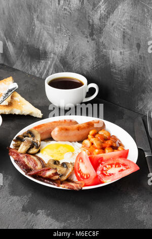 Traditionelles Englisches Frühstück - Speck, Würstchen, Spiegeleier, Tomaten, Bohnen, Pilze, Toast mit Butter - In weißen Teller und Kaffee auf einem dunklen zurück Stockfoto