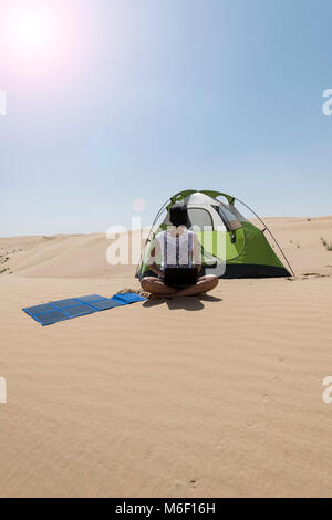 Eine Frau sitzt neben ihrem Zelt in der Wüste, mit einem Laptop, der von flexiblen Solarpaneelen aufgeladen wird, nachhaltiges Wohnen und erneuerbare Energie, vertikale Sicht, heiß Stockfoto