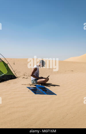 Frau, die ihren Laptop mit flexiblen Solarpaneelen in der Wüste neben ihrem Campingzelt auflädt, vertikale Ansicht mit Kopierraum, Nachhaltigkeitsthema Stockfoto