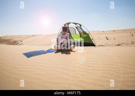 Frau, die ihren Laptop mit flexiblen Solarpaneelen in der Wüste neben ihrem Campingzelt auflädt, Selbstvertrauen und Nachhaltigkeit in einem abgelegenen Bereich Stockfoto