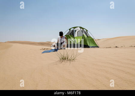 Eine Erwachsene Frau lädt ihren Laptop mit einem flexiblen Solarpanel in der Wüste auf, mit ihrem Campingzelt im Hintergrund, Selbstvertrauen und Nachhaltigkeit Stockfoto