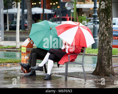 Akteure in Kostümen eine Pause auf einer öffentlichen Bank unter bunten Sonnenschirmen an einem regnerischen nasser Sommer Tag in Wien Stockfoto