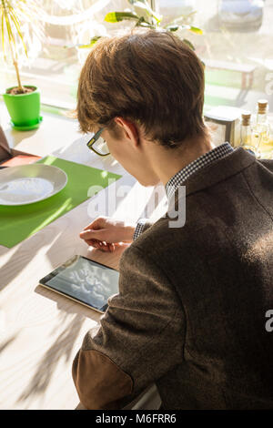 Hübscher junger Mann mit Tablet-PC im Cafe. Männliche Teenager in Tweed jaclet und Brillen verwendet die tragbaren Computer für das Mittagessen im Cafe warten Stockfoto