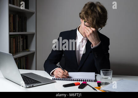 Erschöpft Mitarbeiter oder Student in offiziellen Anzug der späten Stunden bei modernen Schreibtisch Frist für die Zuordnung oder home Aufgabe zu erfüllen. Stockfoto