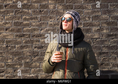 Junger Mann im Winter Kleidung hält Tasse Kaffee. Gut aussehender Teenager männliche Person in Sonnenbrille und Strickmütze Getränke heiße Getränke auf helle, sonnige Winter Stockfoto