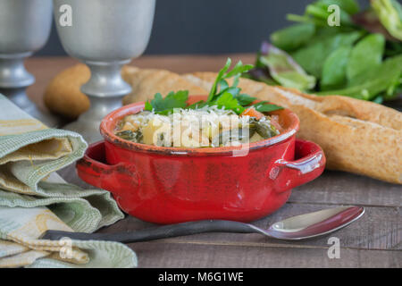 Herzhaftes aus weißen Bohnen und Gemüse Suppe Eintopf serviert in einem roten Topf mit einem Laib Weizen französischem Brot. Essen Szene, hausgemachte Güte. Stockfoto