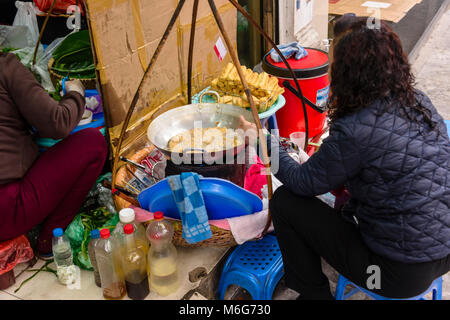 Eine Frau richtet ihre Street Food pop-up Restaurant auf einem Fußweg in Hanoi, Vietnam. Sie trägt alles, was Sie benötigt, zu kochen und zu Kunden, einschließlich Kunststoff Stühle und Tische, Zutaten und ein Herd in Körben, die Sie trägt mit einem Bambusstab über ihre Schulter dienen. Stockfoto