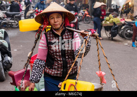 Eine Frau trägt alles, was Sie benötigt, zu kochen und zu bedienen Kunden, einschließlich Kunststoff Stühle und Tische, Zutaten und ein Herd in Körbe mit einem Bambusstab über ihre Schulter in Hanoi, Vietnam Stockfoto