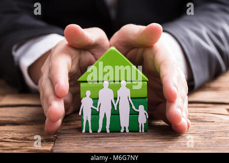 Der Unternehmer schützende Hand Haus Modell und Familie Papier schneiden Sie auf hölzernen Schreibtisch Stockfoto