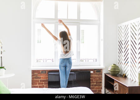 Frau Stretching im Schlafzimmer am Fenster nach dem Aufwachen. Rückansicht, Modell im Schlafzimmer schaut aus dem Fenster in seine Arme über den Kopf hob. Stockfoto