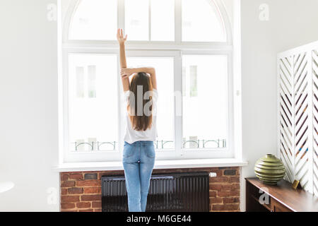 Frau Stretching im Schlafzimmer am Fenster nach dem Aufwachen. Rückansicht, Modell im Schlafzimmer schaut aus dem Fenster in seine Arme über den Kopf hob. Stockfoto