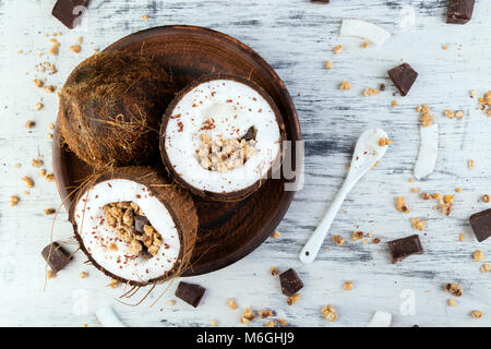 Gesundes Frühstück in Coconut Bug auf weißem Hintergrund. Joghurt in Kokosnuss Schale mit Kokosraspeln, Schokolade und Müsli. Ansicht von oben, flach, Overhead Stockfoto