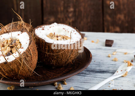 Gesundes Frühstück in Coconut Bug auf weißem Hintergrund. Joghurt in Kokosnuss Schale mit Kokosraspeln, Schokolade und Müsli. Ansicht von oben, flach, Overhead Stockfoto