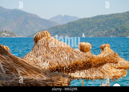 Strohschirme stehen im Vordergrund an einem sonnigen Strand mit klarem blauem Meer und Bergketten im Hintergrund Stockfoto