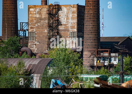 Verlassener Industriekomplex mit hohen, verwitterten Schornsteinen, die über den baufälligen Gebäuden vor einem klaren blauen Himmel thronen Stockfoto