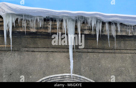 Feder Eiszapfen hängen von Schnee bedeckte Dach auf einem blauen Himmel Hintergrund. Eiszapfen sind sehr gefährlich für das Leben/gefährliche saisonale Leute Wetter Stockfoto