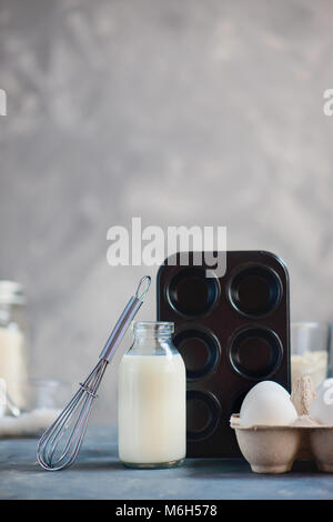 Backen Werkzeuge und Zutaten auf einen konkreten Hintergrund. High key Essen Fotografie mit einem Muffin tin, Schneebesen, eine Flasche Milch und einen Karton Eier. Kopieren Stockfoto