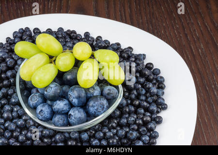 Blaubeeren und Trauben auf Tisch. Vaccinium myrtillus. In der Nähe von weißen Teller mit frischen Früchten und Glas Schüssel auf Holz- Hintergrund. Stockfoto