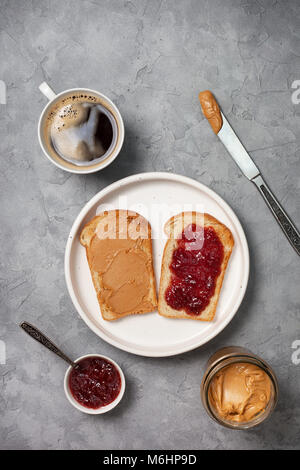 Zwei Toast mit Erdnussbutter und Marmelade, eine Tasse Kaffee auf grauem Beton Hintergrund. Ansicht von oben Stockfoto