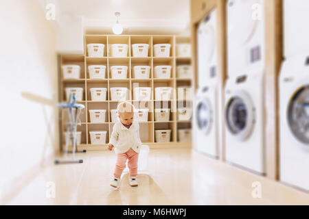 Das Baby befindet sich in der Waschküche auf dem Hintergrund von Regalen und Waschmaschinen mit Lächeln Stockfoto
