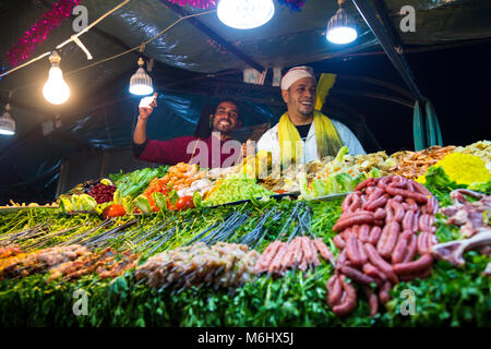 Köche lächelnd an einer Garküche voll mit frischem Gemüse und Fleisch am Spieß in Jemaa el-Fnaa Markt, Marrakesch, Marokko Stockfoto