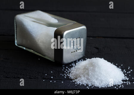 Glas Salt Shaker auf Schwarz rustikalen Tisch und Haufen von Salzkristallen mit Platz für Text im Hintergrund Stockfoto