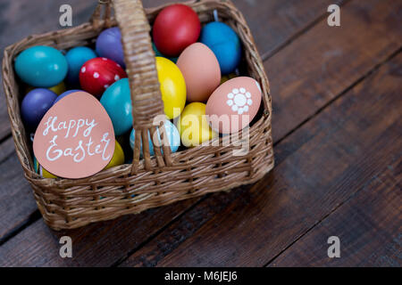 Hintergrundbild mit Weidenkorb mit bunten Ostereier in auf rauen Holztisch mit Frohe Ostern Hinweis Stockfoto