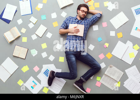 Junger Mann auf grauem Hintergrund mit Papieren und fest schlafen mit Buch isoliert Stockfoto