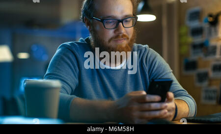 Junger bärtiger Mann Typen Nachricht auf seinem Smartphone. Er sitzt am Schreibtisch und lächelt. Stockfoto