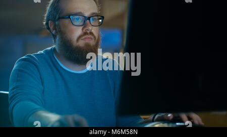 Schwerpunkt junge Mann arbeitet auf einem Personal Computer. Er trägt eine Brille und Bart, er arbeitet bis spät in die Nacht im Büro Dachboden.