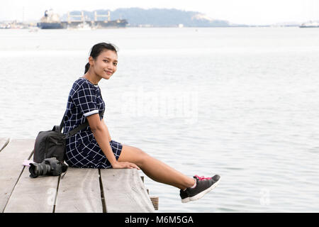 Junge Frau mit Kamera sitzen auf einem hölzernen Pier über das Meer. Asiatische Touristen entspannen am Meer, Penang, Malaysia. Stockfoto