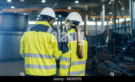 Ansicht von hinten erschossen von Männlichen und Weiblichen Wirtschaftsingenieure in Diskussion und Verwendung Laptop während man durch Heavy Industry Manufacturing Factory. Stockfoto