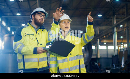 Männliche und weibliche Ingenieure in Diskussion und Verwendung Laptop während man durch Heavy Industry Manufacturing Factory. Sie harte Hüte tragen Stockfoto