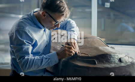 Senior Automotive Designer mit Rechen formt futuristischen Auto Modell aus Plastilin Ton. Er arbeitet in einem speziellen Studio In einem großen Auto Fabrik befindet. Stockfoto