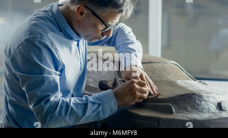 Senior Automotive Designer mit Rechen formt futuristischen Auto Modell aus Plastilin Ton. Er arbeitet in einem speziellen Studio In einem großen Auto Fabrik befindet. Stockfoto