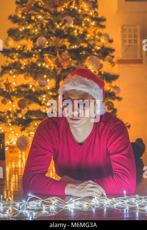 Nachdenklicher Mann mit Santa Hut auf dem Boden liegen und Blick auf Weihnachtsbeleuchtung. Weihnachtsbaum im Hintergrund Stockfoto