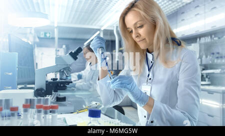 Weibliche Wissenschaftler verwendet Mikropipette Füllung Röhrchen in einem großen modernen Labor. Im Hintergrund arbeiten die Wissenschaftler. Stockfoto