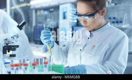 Weibliche Wissenschaftler verwendet Mikropipette Füllung Röhrchen in einem großen modernen Labor. Im Hintergrund arbeiten die Wissenschaftler. Stockfoto