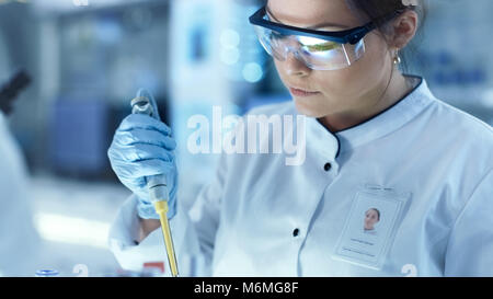 Weibliche Wissenschaftler verwendet Mikropipette Füllung Röhrchen in einem großen modernen Labor. Im Hintergrund arbeiten die Wissenschaftler. Stockfoto