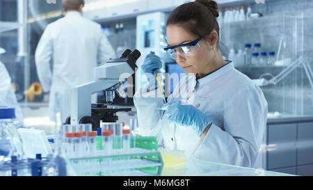 Weibliche Wissenschaftler verwendet Mikropipette Füllung Röhrchen in einem großen modernen Labor. Im Hintergrund arbeiten die Wissenschaftler. Stockfoto