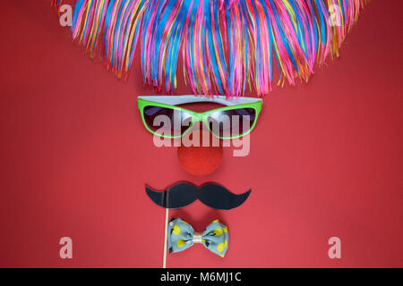 Flach der Clown aus bunten Perücke, Sonnenbrille, rote Nase, Papier prop Schnurrbart und Bow Tie gegen roten Hintergrund. Minimalen Begriff. Stockfoto