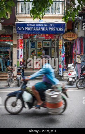 HANOI, VIETNAM - 14. Juli: Ein Mann transportiert ein Faß auf ein Moped vorbei an einem alten Propaganda Poster Shop in der Altstadt von Hanoi am 14 Juli, 2017 in Han Stockfoto