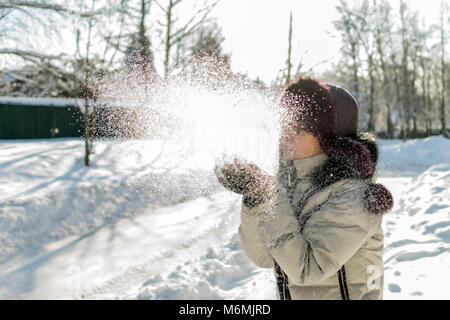 Hält eine Frau eine Handvoll Schnee in den Händen und bläst. Winter im Wald. Die sonne funkelt. Tag, Russland. Stockfoto