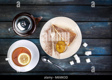 Stück Käsekuchen mit Marmelade und Tee Wasserkocher mit Zitrone auf einem farbigen Hintergrund aus Holz. Ansicht von oben Stockfoto