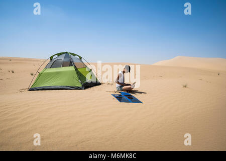 Frau, die ihren Laptop mit flexiblen Solarpaneelen in der Wüste neben ihrem Campingzelt auflädt, Selbstvertrauen und Nachhaltigkeit, Kopierraum Stockfoto