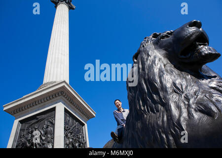 Ein junges Mädchen sitzt auf einer der Löwenstatuen am Fuß der Nelsons-Säule in der Hauptstadt von Großbritannien, London. Beliebt bei Touristen, Urlaubern und Menschen im Urlaub genießen sie es, auf dem Rücken der riesigen Bronzeskulpturen zu sitzen und ihre Fotos zu machen. Stockfoto