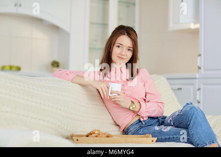 Ein junges Mädchen mit einem Becher heißen Getränk sitzt im Zimmer. Stockfoto