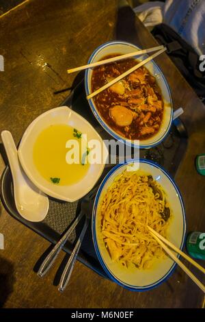 Oben Blick auf drei Teller mit Essen, Nudeln, Hähnchen und Suppe über einen hölzernen Tisch in einem Restaurant mit choopsticks und Holzlöffel in einem Restaurant inSentosa Island, Singapur Stockfoto