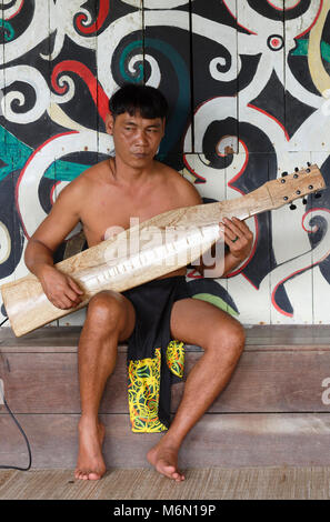 Native Mann spielt traditionelle Musik auf einem elektrischen Sape in der Orang East Langhaus, vor dem Baum des Lebens Wandbild im Sarawak Cultural Village Stockfoto