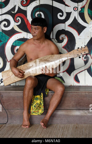 Native Mann spielt traditionelle Musik auf einem elektrischen Sape in der Orang East Langhaus, vor dem Baum des Lebens Wandbild im Sarawak Cultural Village Stockfoto